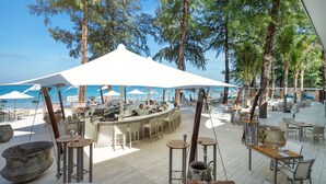 Eine Außenbar mit Sitzgelegenheiten und Sonnenschirmen mit Blick auf einen Strand, an dem Boote und Palmen zu sehen sind.