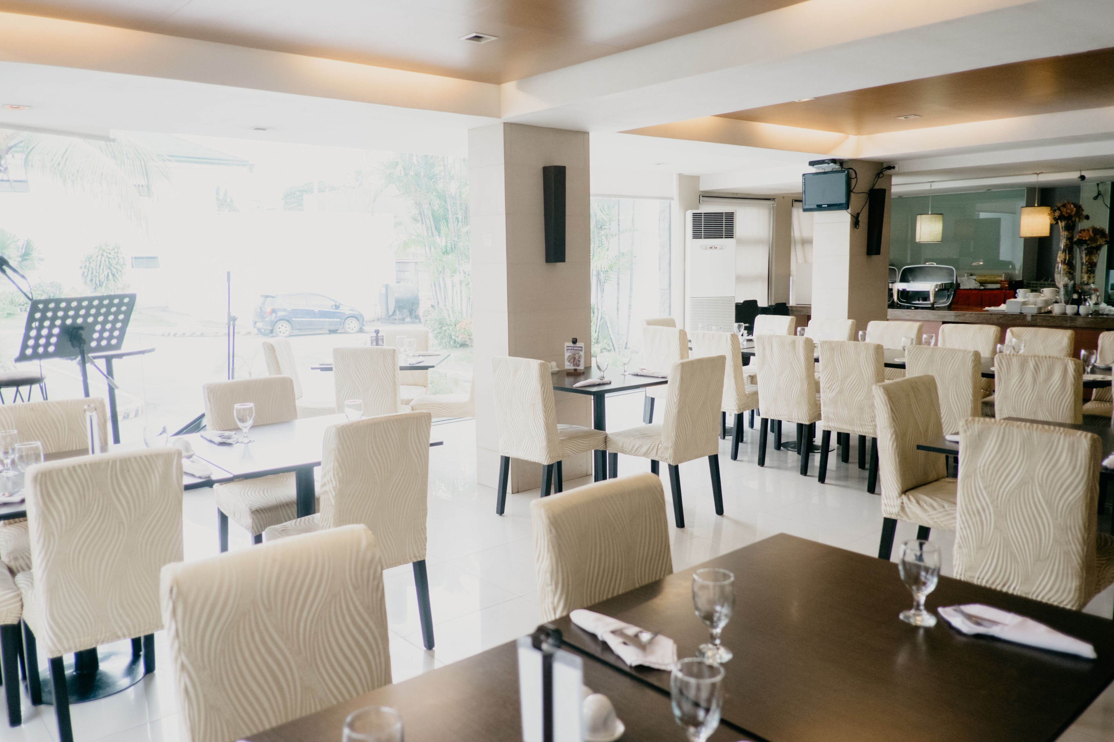 A modern dining area with white upholstered chairs and dark tables, large windows offering a view of the street outside.