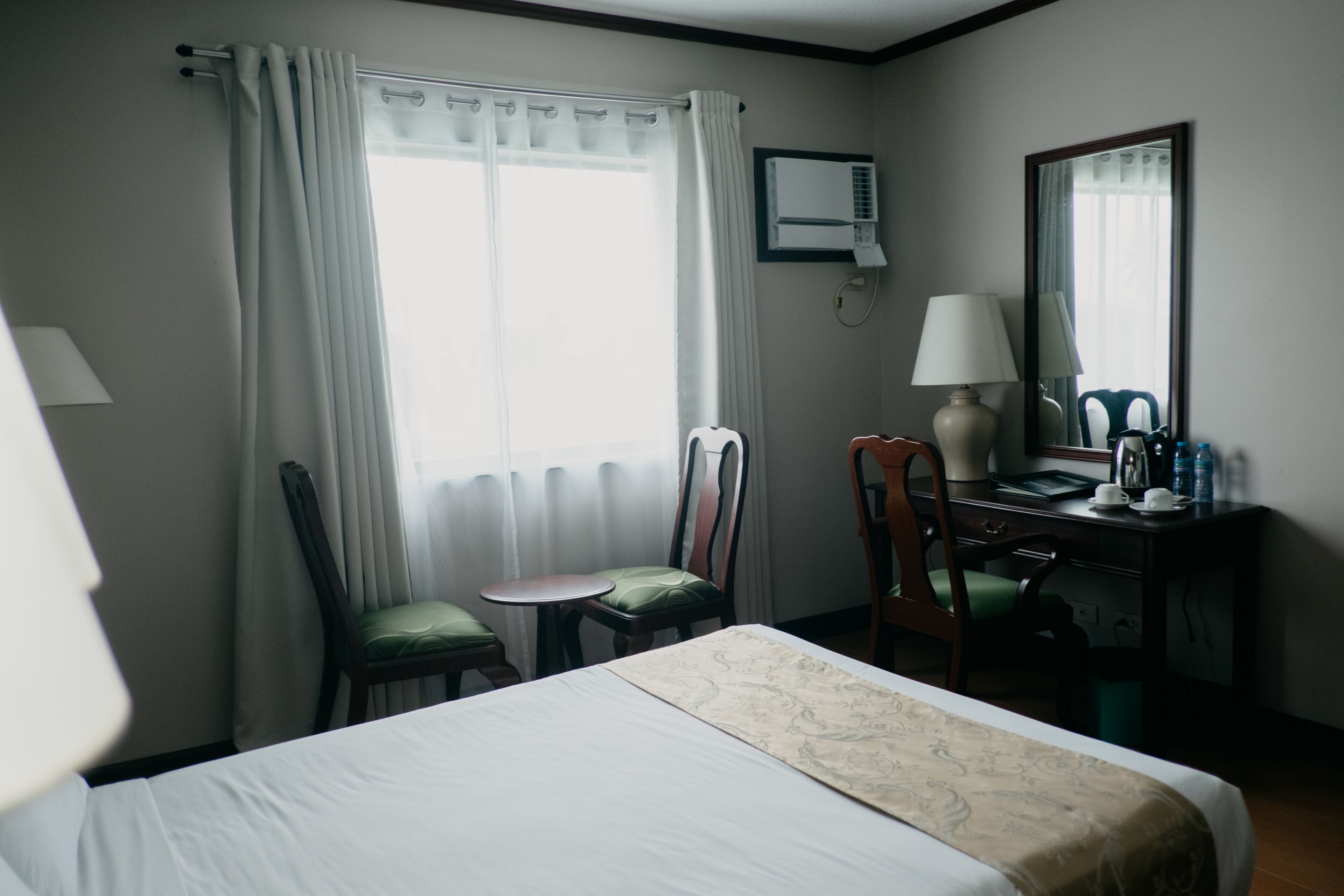 Una habitación de hotel con cama, escritorio, silla, mesita, espejo y unidad de aire acondicionado.