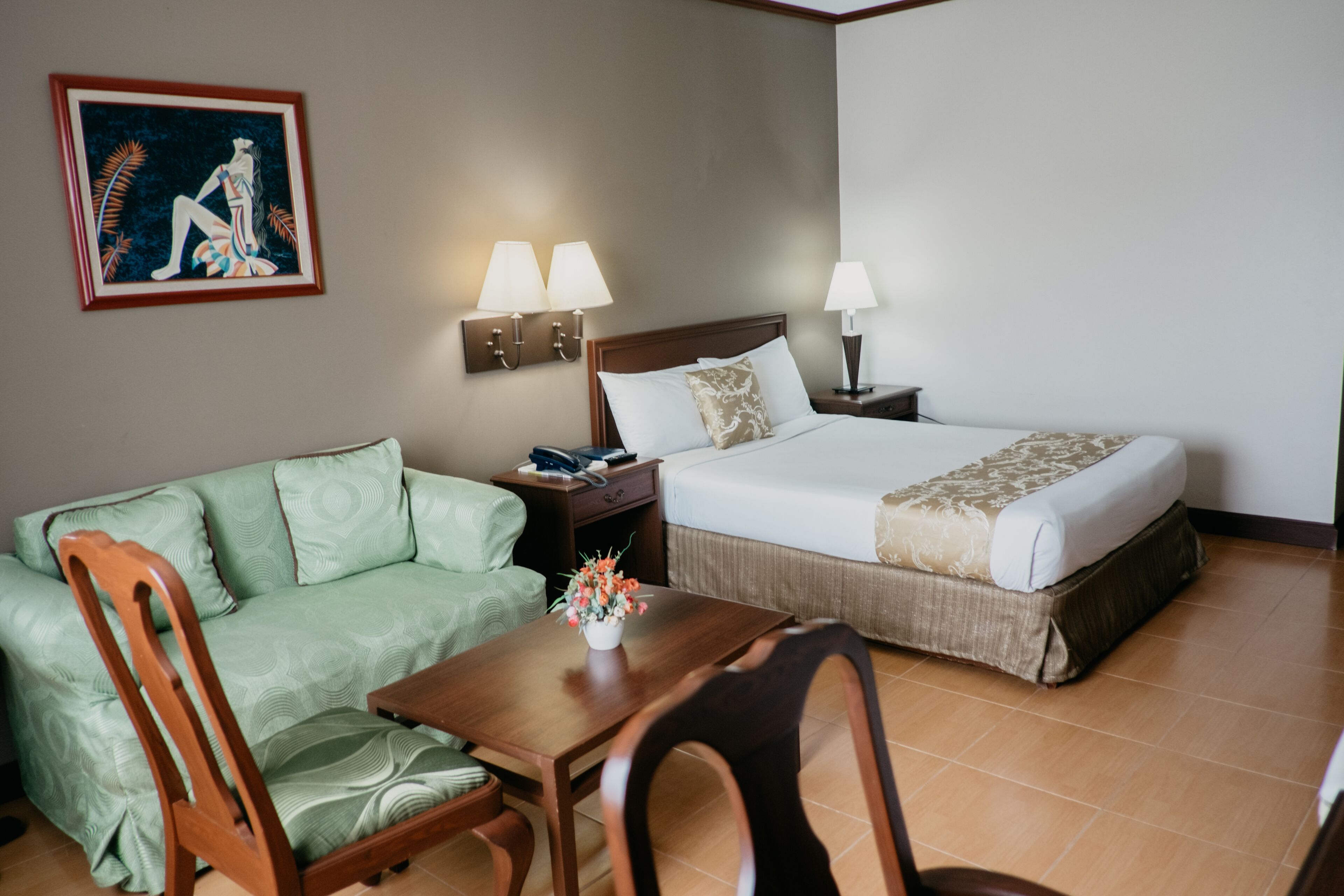 Una habitación de hotel con una cama, un sofá verde, una mesa de madera con un jarrón de flores y un cuadro en la pared.