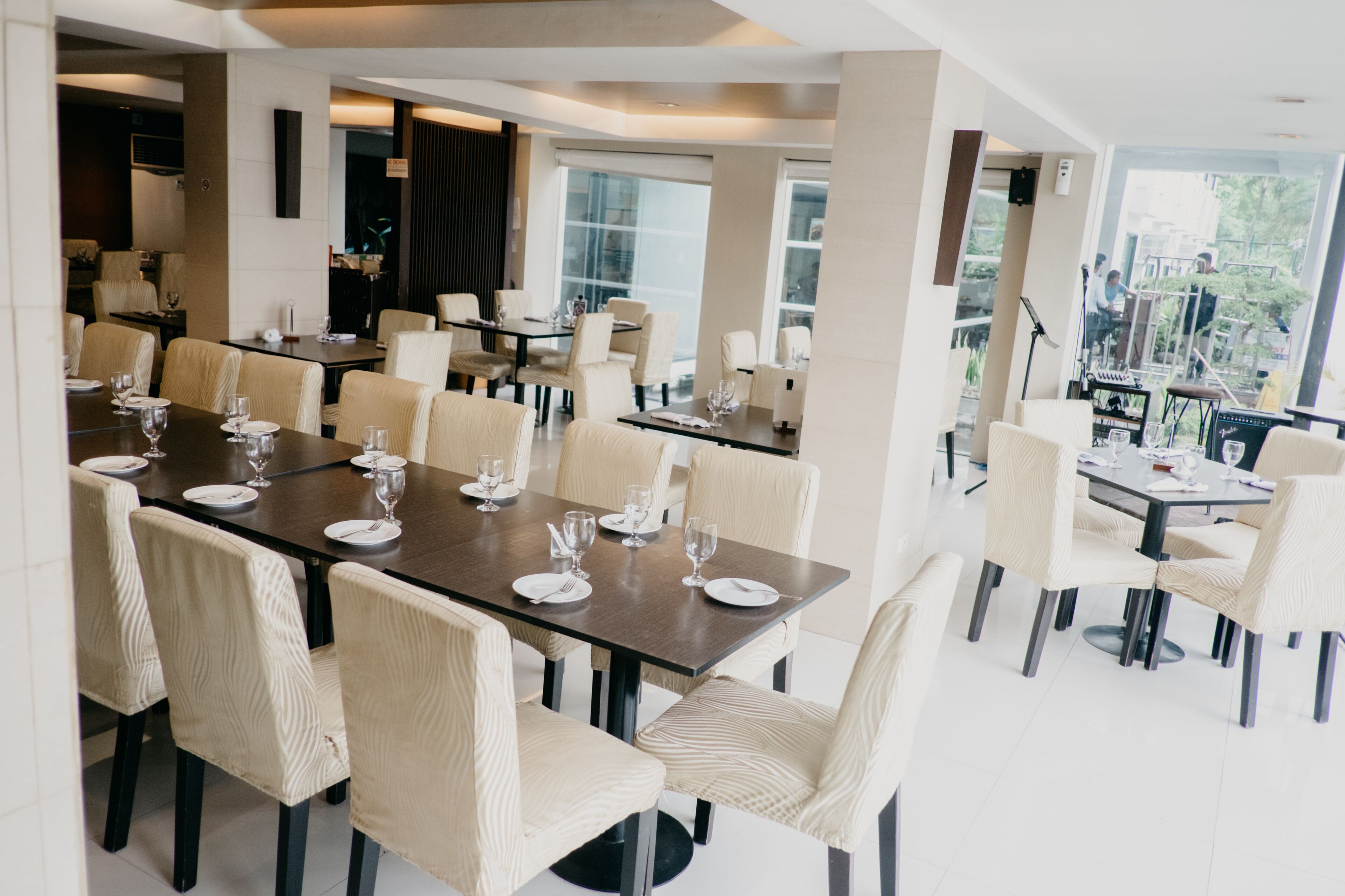 A modern dining area with multiple tables and chairs, large windows, and a view of the outdoors.