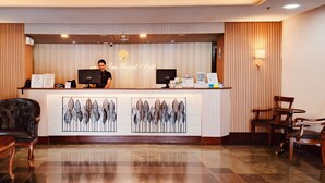 Zona de recepción de hotel con un mostrador, una computadora, un teléfono y una máquina de café. Hay dos sillas y una mesa en primer plano.