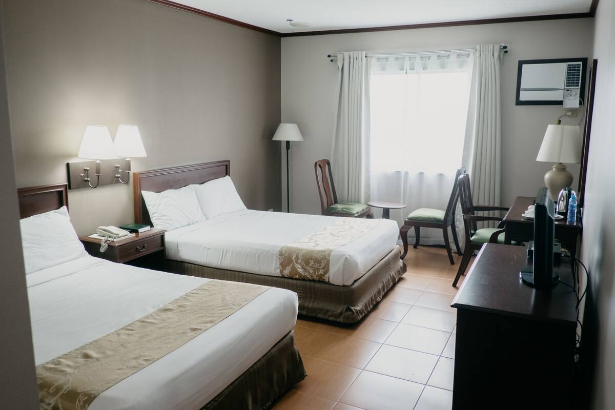 Ein Hotelzimmer mit zwei Betten, einem Schreibtisch, einem Stuhl, einer Lampe und einem Fenster mit Vorhängen.