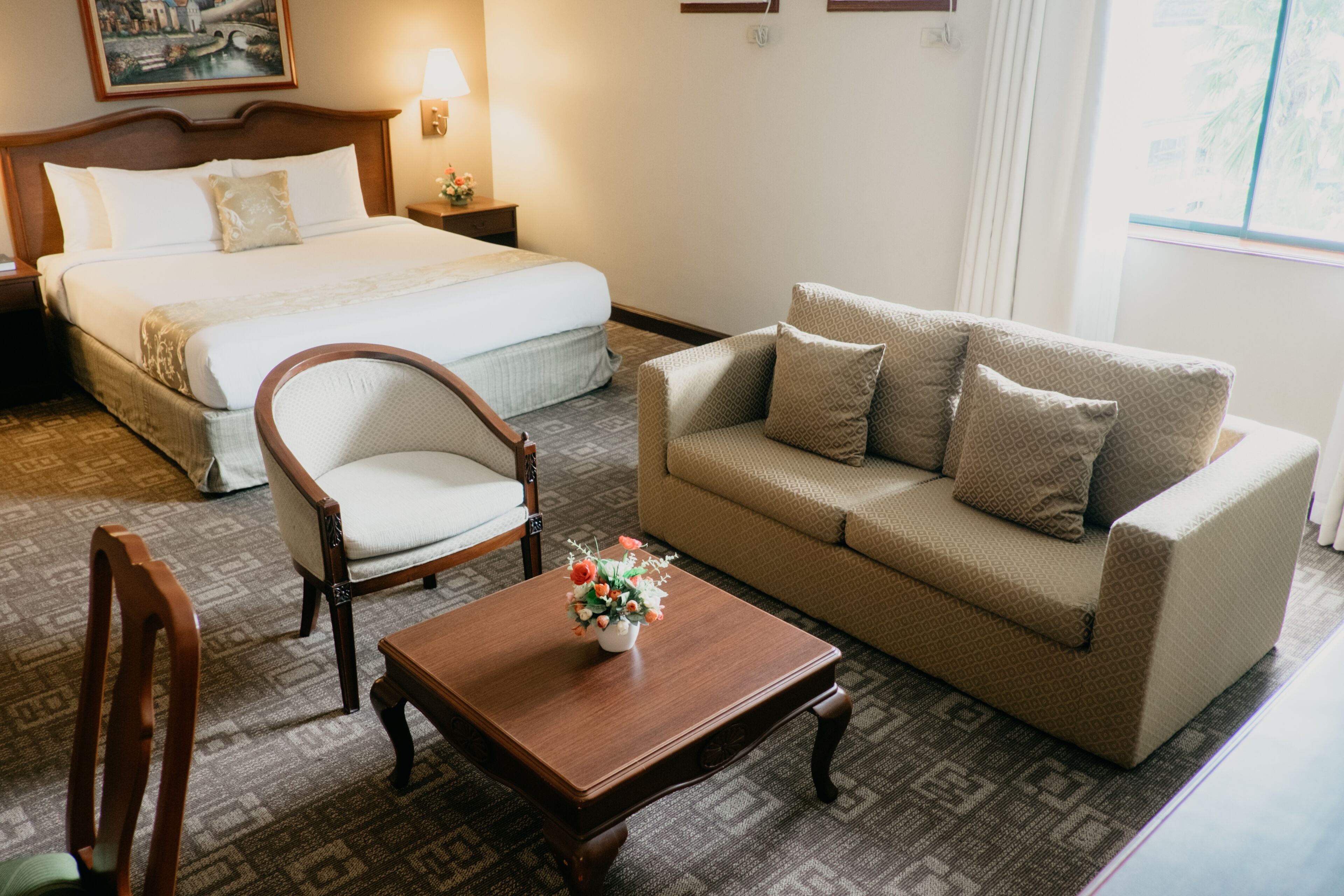 A hotel room with a bed, a sofa, a wooden chair, a small table with a vase of flowers, and a nightstand with a lamp.