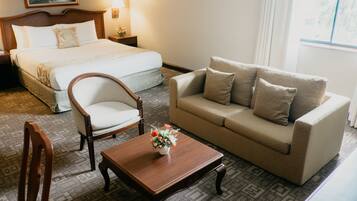 A hotel room with a bed, a sofa, a wooden chair, a small table with a vase of flowers, and a nightstand with a lamp.