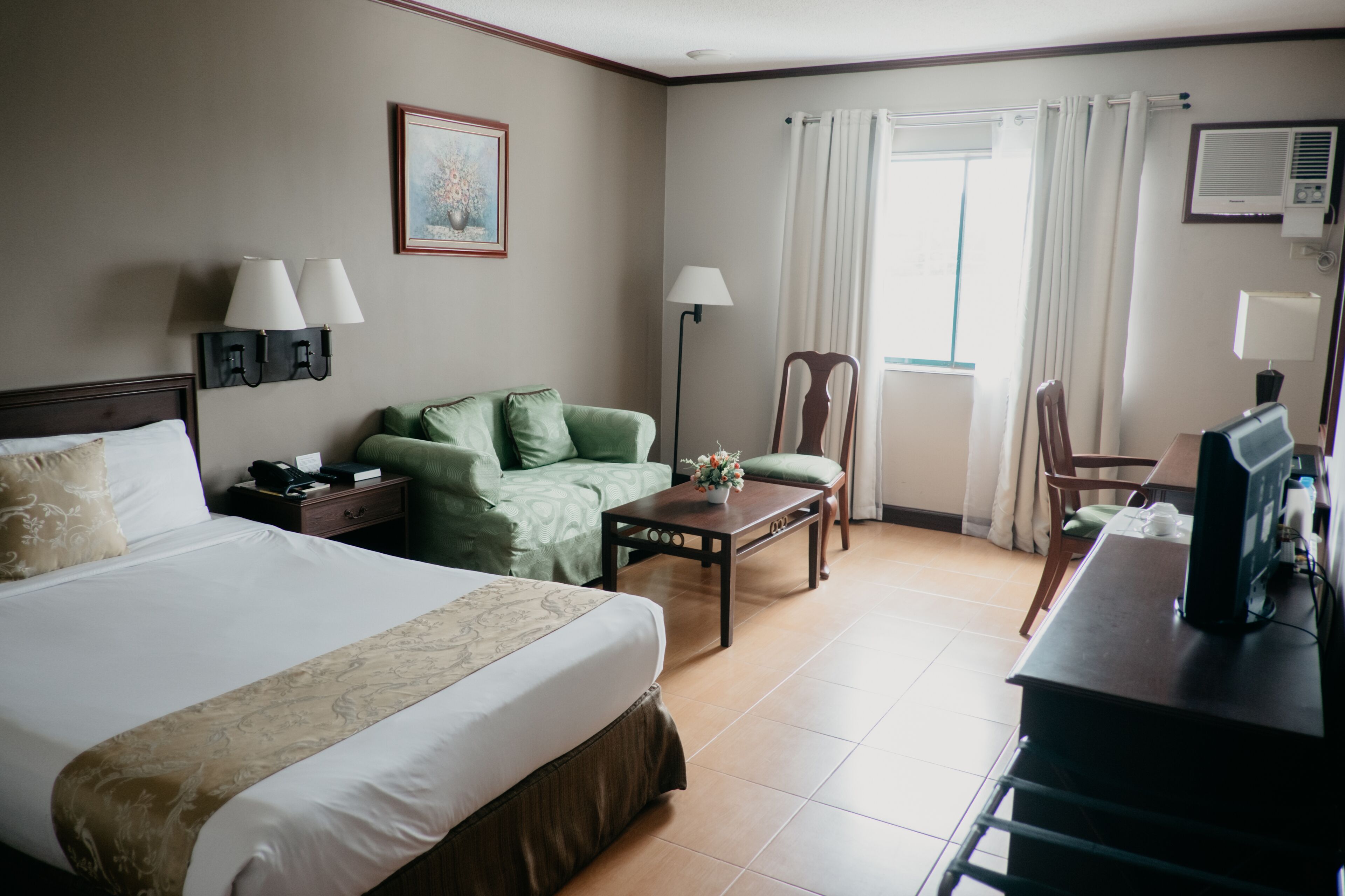 A hotel room with a large bed, a green sofa, a wooden coffee table, a desk with a computer, and a window with curtains.