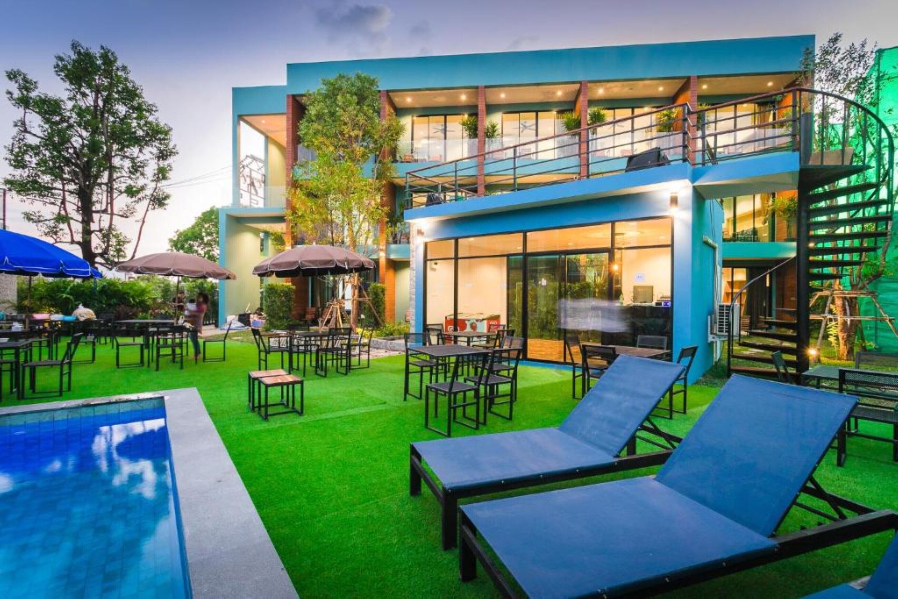 Un área de piscina moderna al aire libre con sillones de descanso, un comedor y un edificio de dos pisos con ventanas grandes y una escalera.