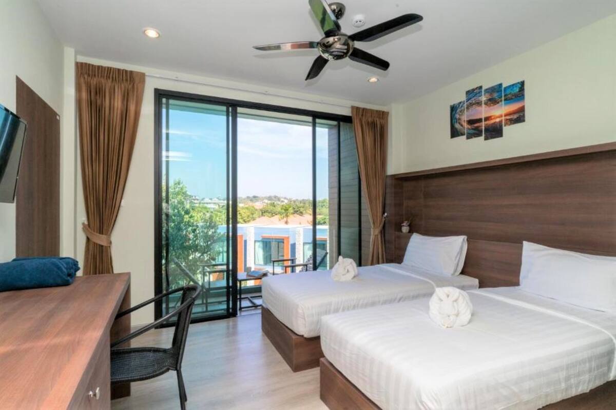 Ein Hotelzimmer mit zwei Betten, einem Schreibtisch und einem Sessel. Es gibt einen Deckenventilator, große Fenster und einen Blick auf Gebäude.