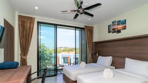 A hotel room with two beds, a desk, and a chair. There is a ceiling fan, large windows, and a view of buildings outside.