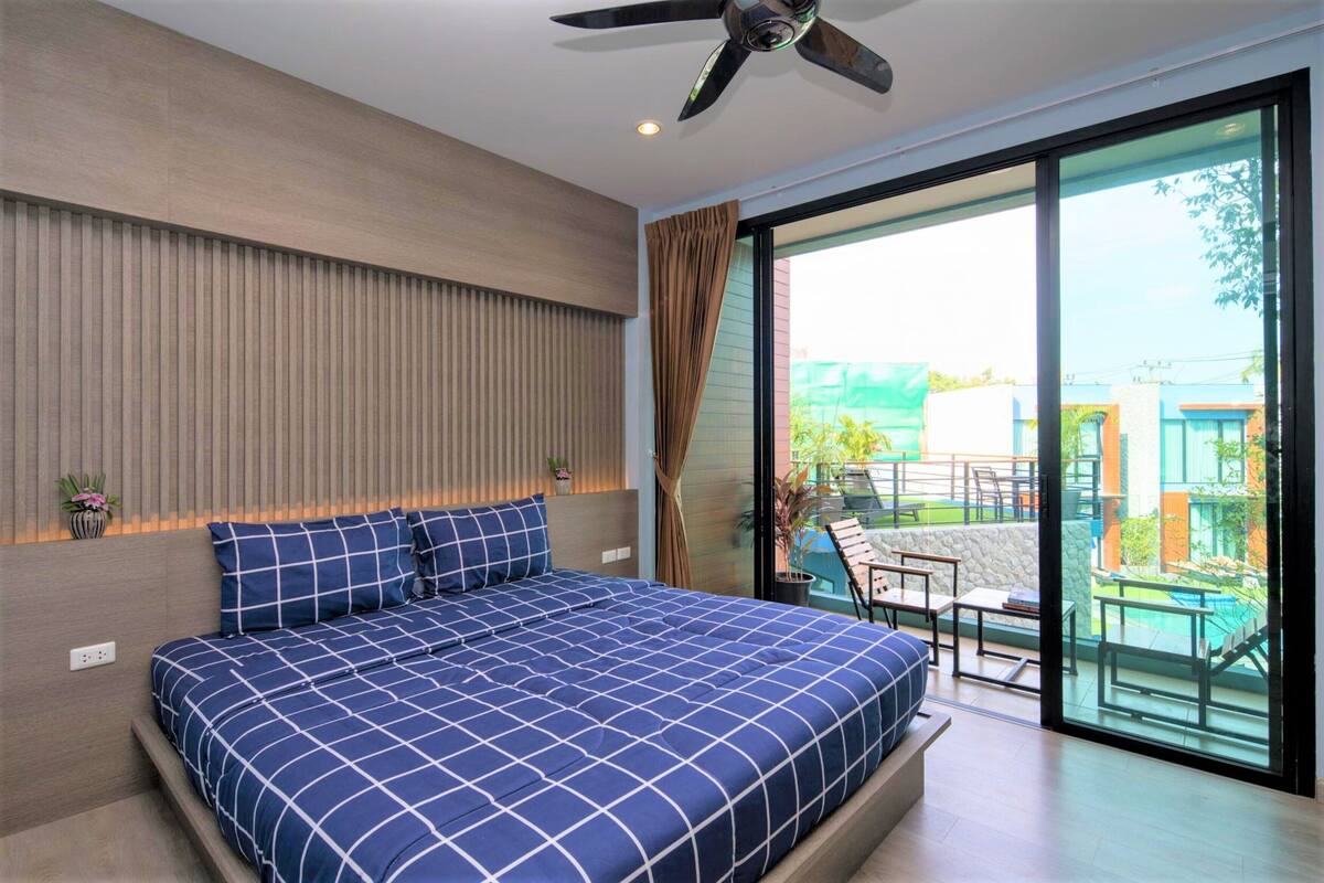 Ein modernes Schlafzimmer mit einem großen Bett, einem Deckenventilator und einer Glastür, die auf einen Balkon mit Sitzmöbeln führt.
