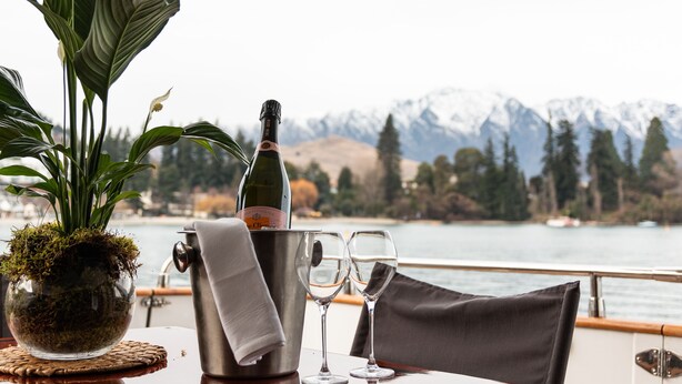 A table with a bottle of champagne, two glasses, a potted plant, and a scenic view of mountains and a lake. - Pacific Jemm - Luxury Super Yacht (Queenstown)