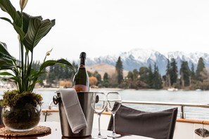 Une table avec une bouteille de champagne, deux verres, une plante en pot et une vue panoramique sur des montagnes et un lac.