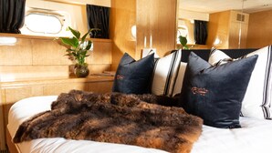 A bed with a fur throw, dark pillows, and a plant on a wooden bedside table. - Pacific Jemm - Luxury Super Yacht (Queenstown)