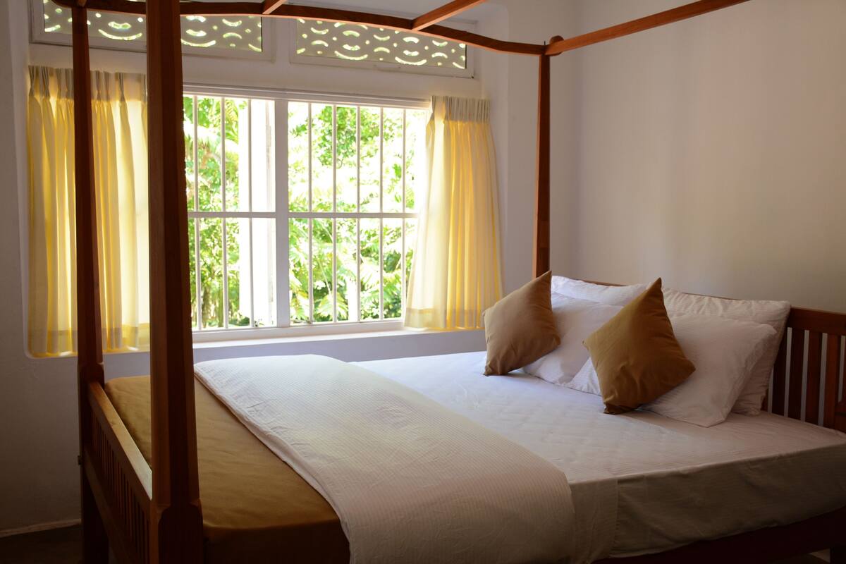 A four-poster bed with a white bedspread and two pillows, a window with curtains, and a view of greenery outside.