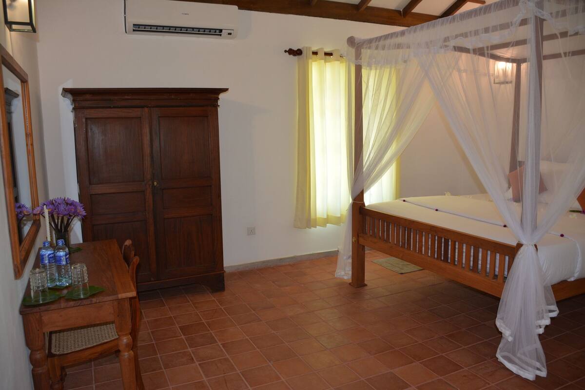 A bedroom with a four-poster bed, a wooden wardrobe, a small table with water bottles, and a window with curtains.