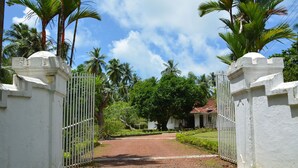 Une entrée fermée par un portail, avec un chemin pavé menant à un bâtiment entouré d’une végétation luxuriante et de palmiers.
