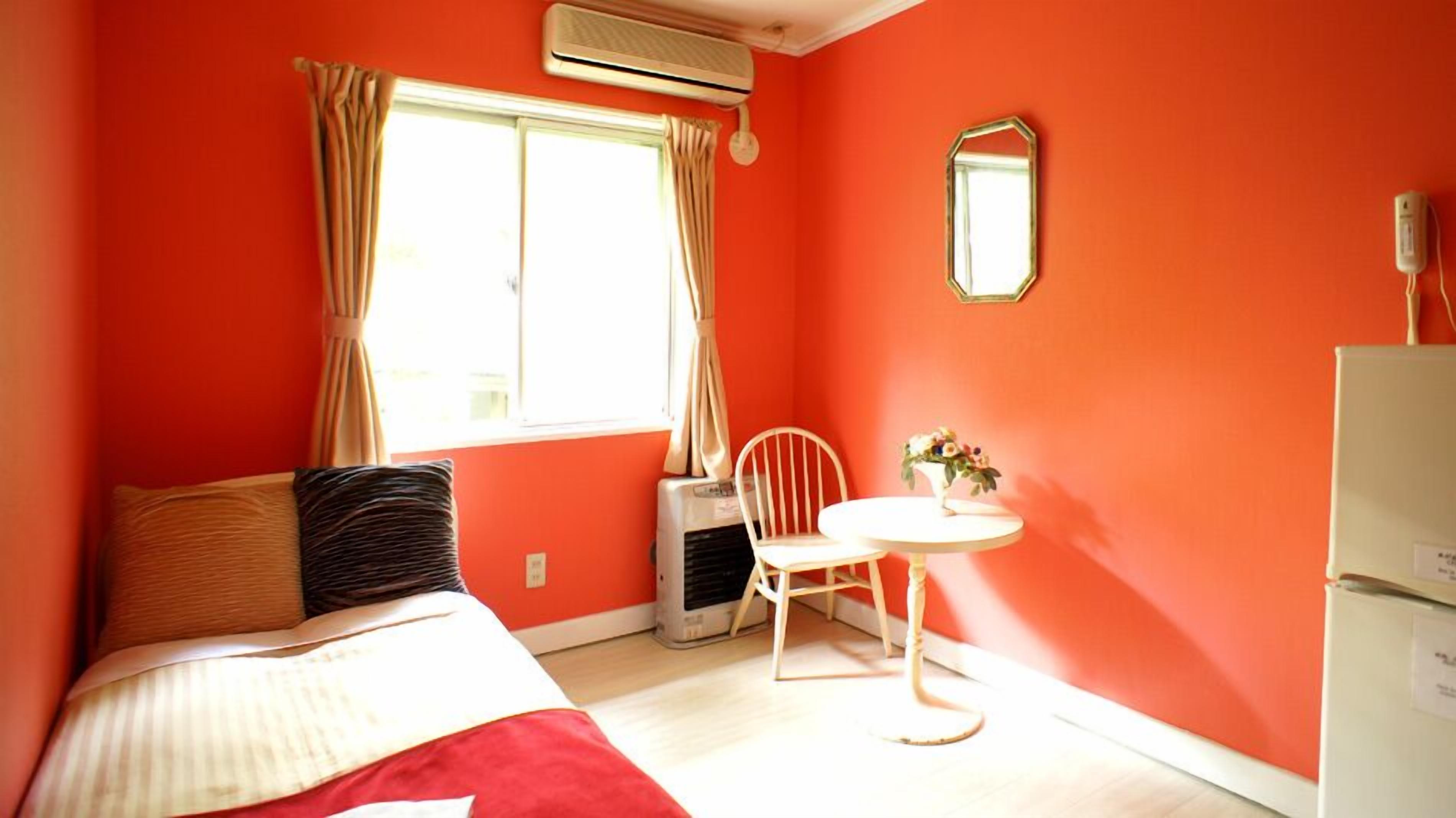 Una habitación con cama, una silla, una mesita, aire acondicionado, un espejo y una ventana con cortinas.