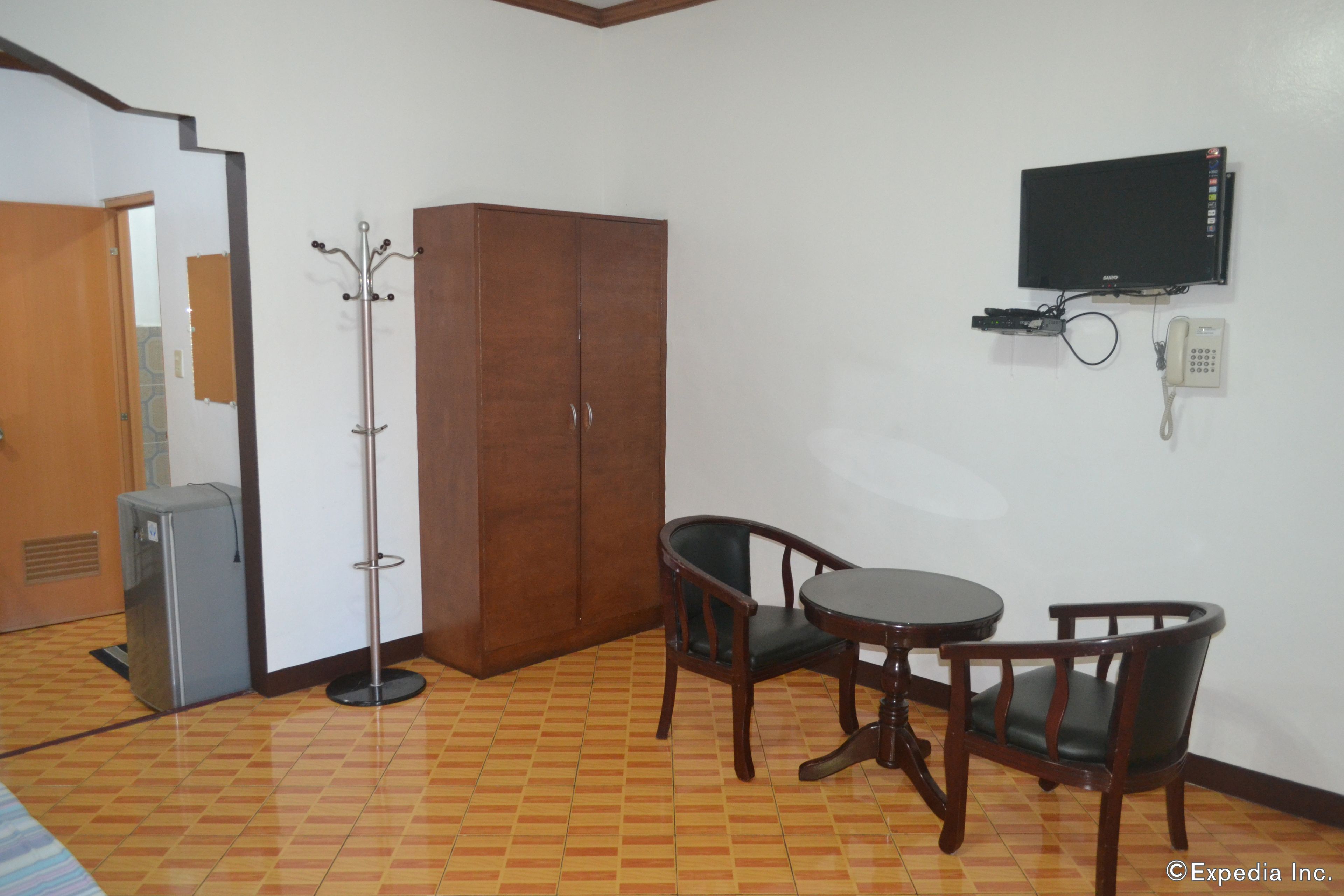 A room with a wooden wardrobe, a coat rack, a television mounted on the wall, a telephone, a small refrigerator, and a dining table with two chairs.
