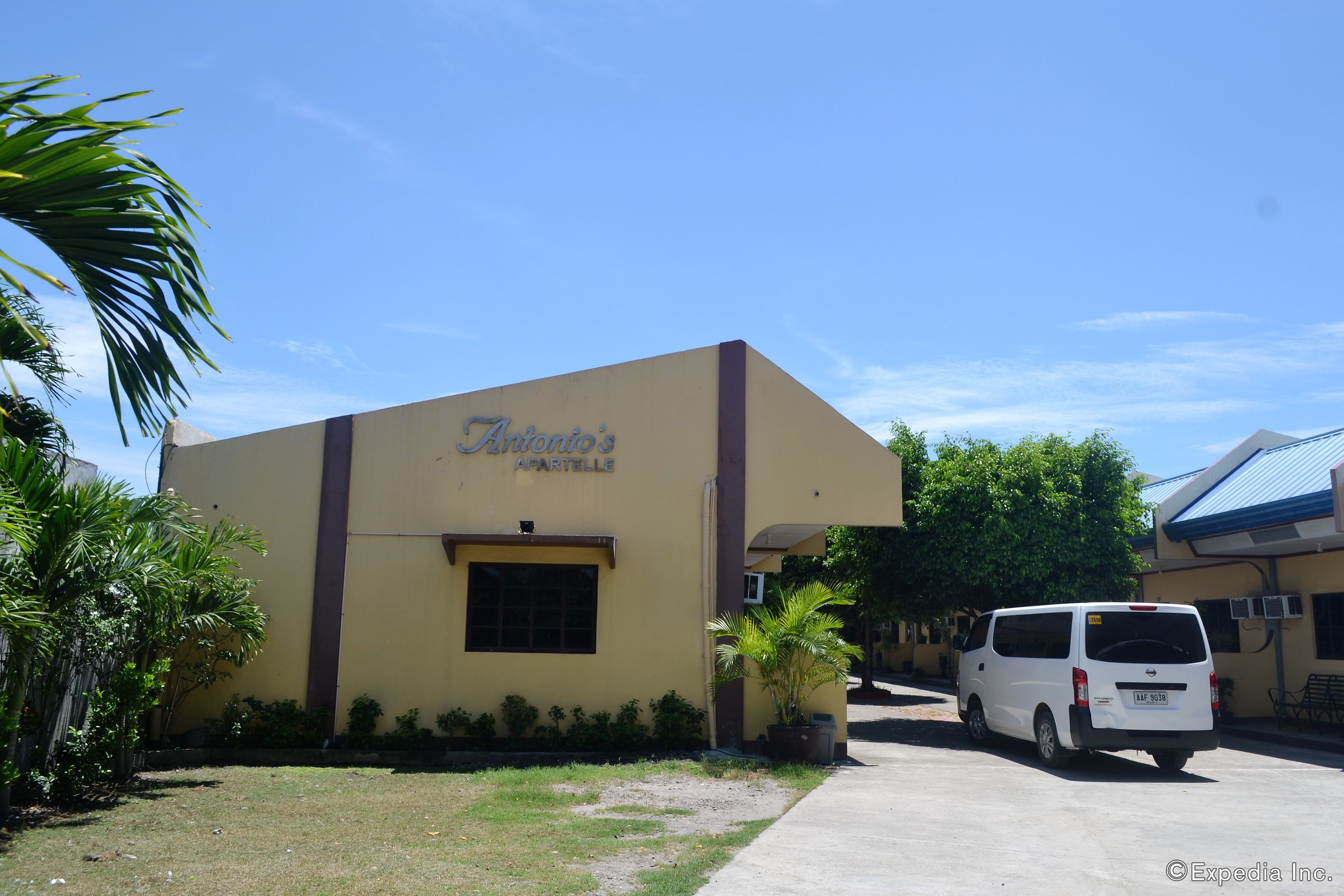 A building with a sign reading "Antonio's Apartelle," a white van parked in front, and a clear blue sky.