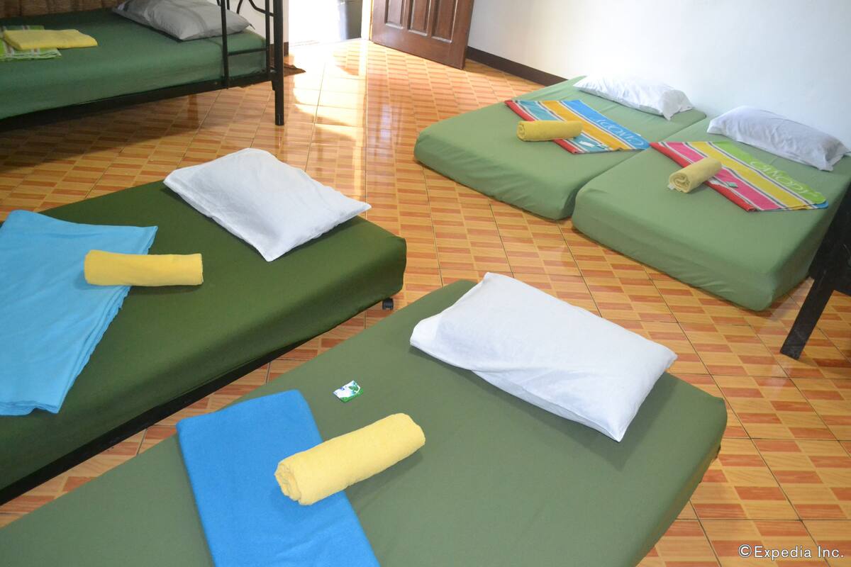 Ein Zimmer mit vier Einzelbetten, jedes mit einer grünen Matratze und weißen Kissen. Auf den Betten liegen Handtücher in Blau, Gelb und Weiß. Der Boden hat ein Schachbrettmuster.
