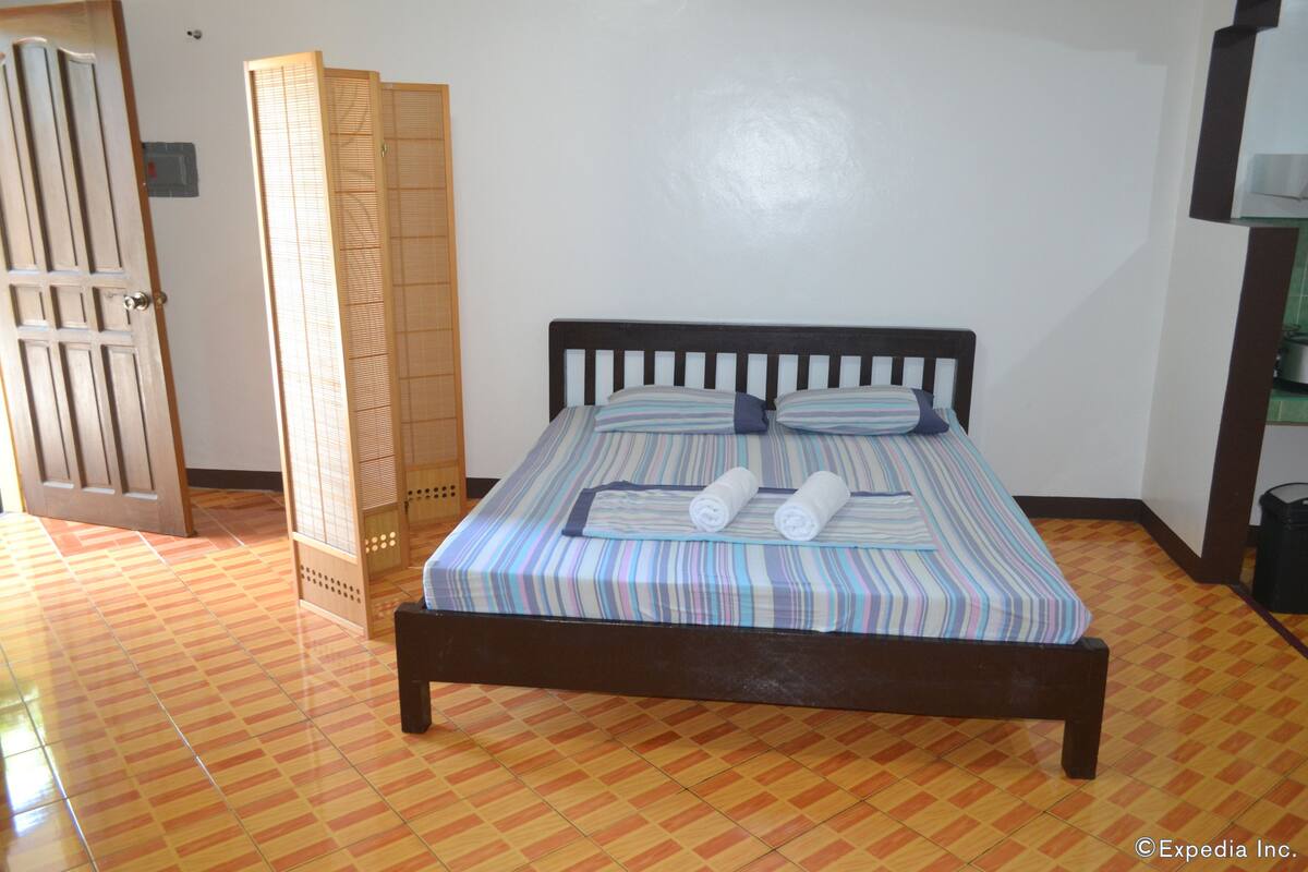 Ein Bett mit gestreifter Bettdecke, zwei gerollte Handtücher, ein hölzernes Kopfende und ein Fliesenboden.