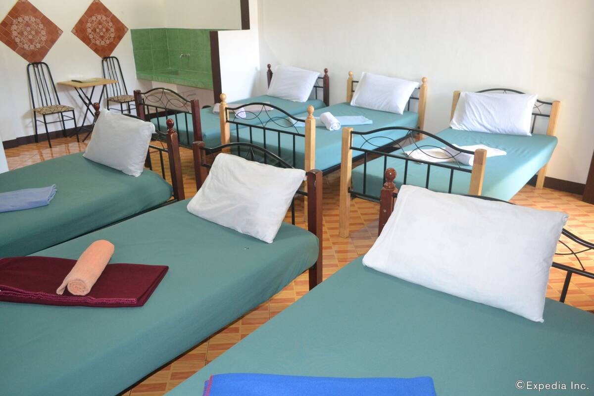 Ein Hotelzimmer mit vier Einzelbetten, jeweils mit Holzbettgestell und grünen Bettlaken. In der Ecke befinden sich zwei Stühle und ein kleiner Tisch.
