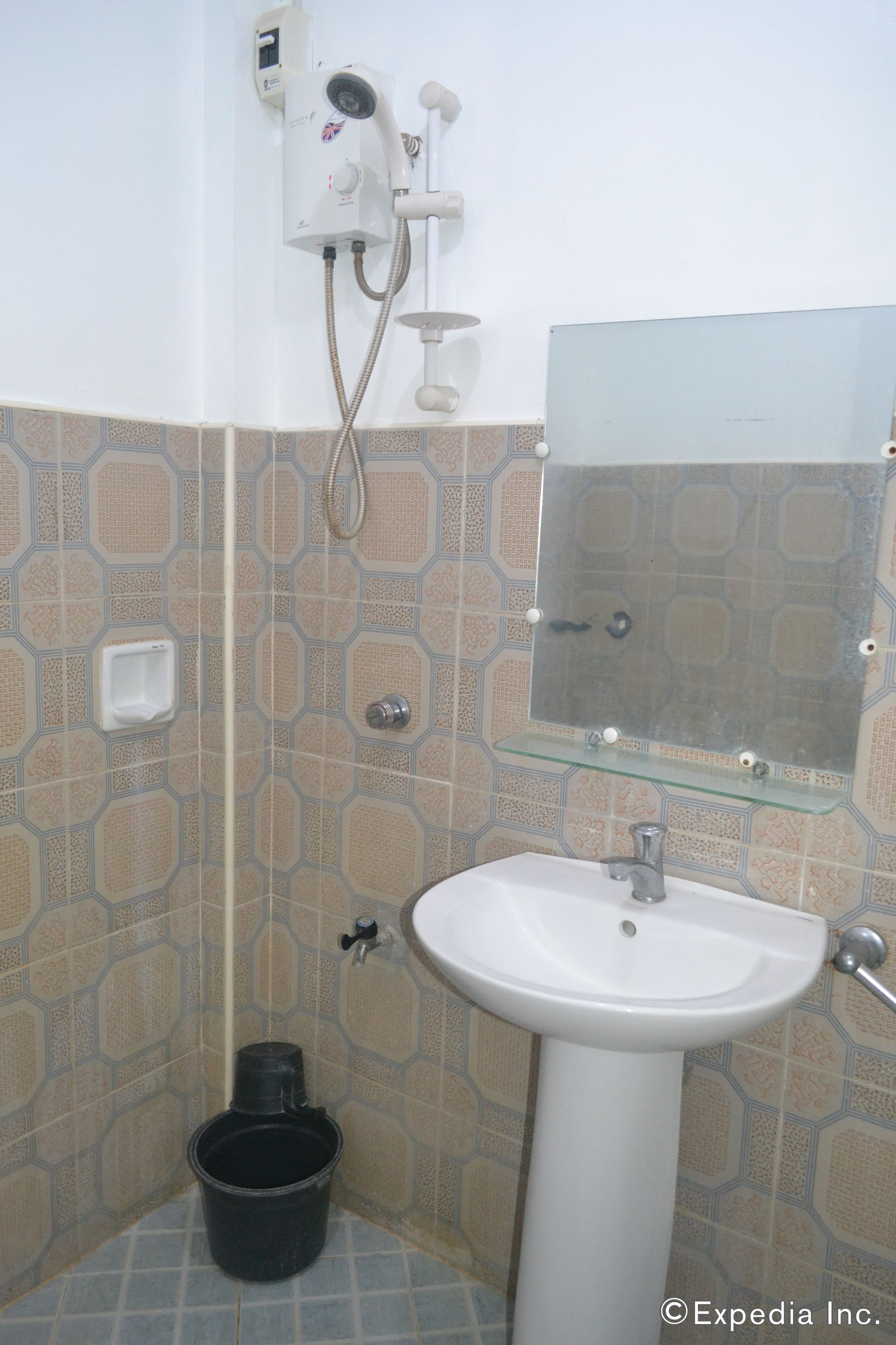 Un baño con un lavamanos blanco, un cabezal de ducha de mano, un estante de vidrio y un cubo negro.