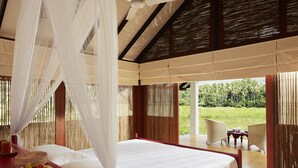 A bedroom with a bed, a bedside table with a book and fruit, a ceiling fan, and a view of a garden. - Jetwing Kaduruketha (Wellawaya)