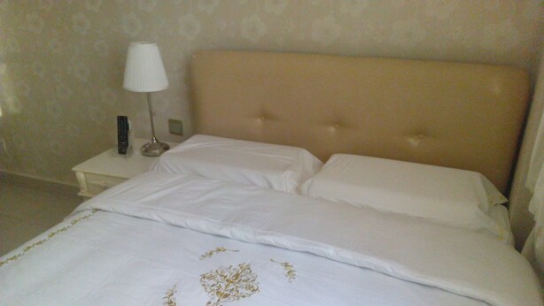Una cama bien hecha con cabecera color beige y ropa de cama blanca, una mesita de noche con lámpara, y una pared con un estampado floral discreto.