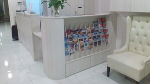 Un área de recepción con un mostrador blanco, una planta en maceta, un sillón blanco y un reloj en la pared.