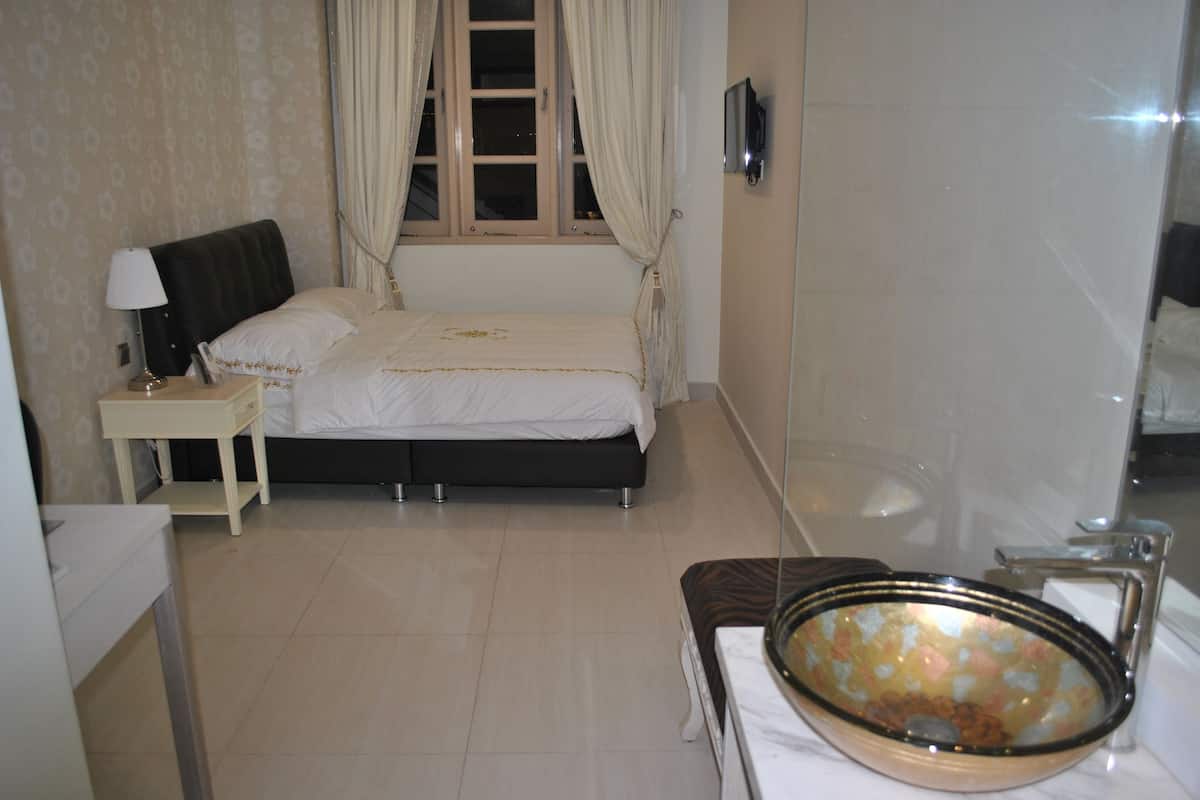 A bedroom with a bed, a nightstand, a bedside table, a window with curtains, and a bathroom with a bathtub and a glass-enclosed shower.