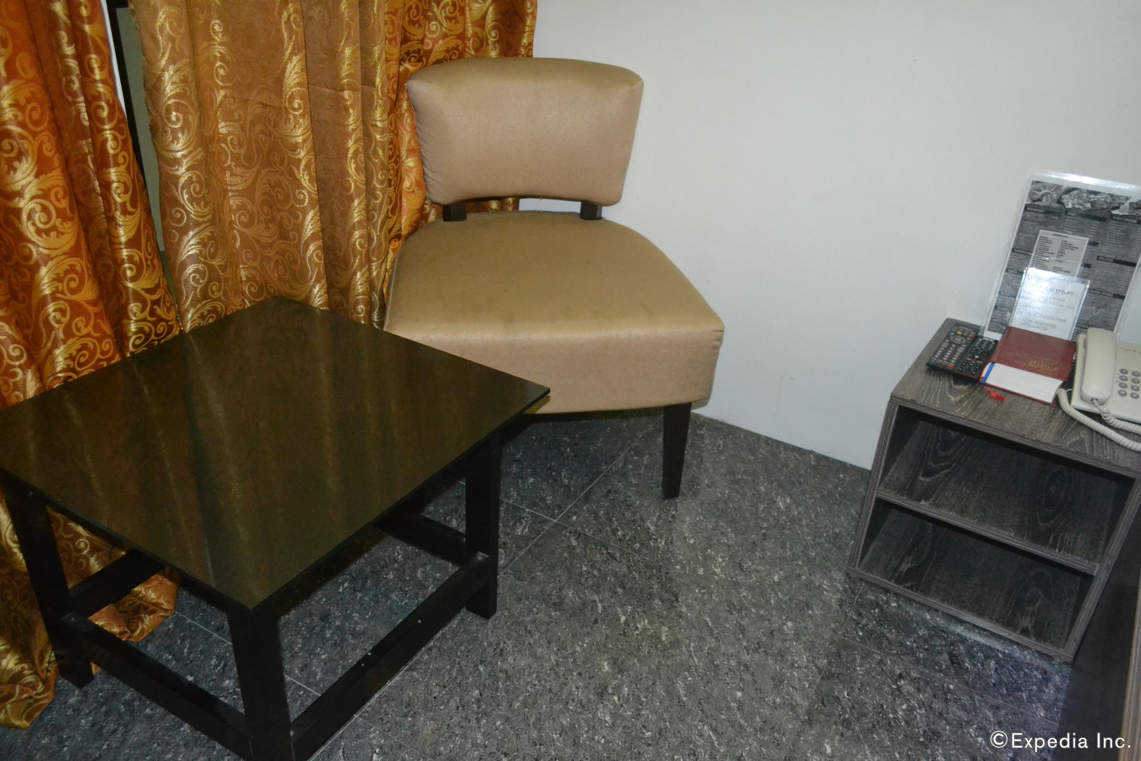 A room with a wooden table, a beige chair, a telephone, and a small shelf with a book and a remote control.