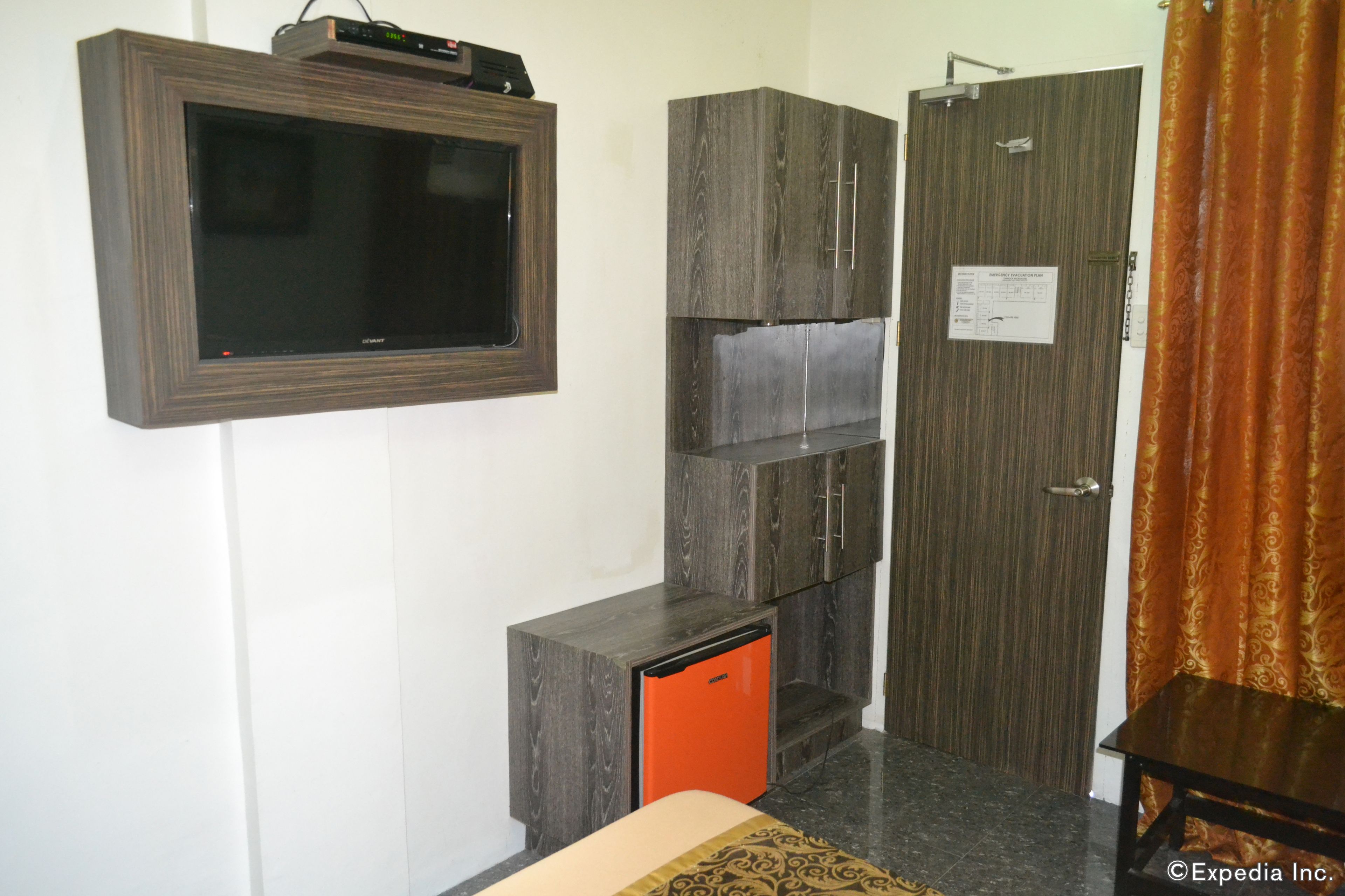 A compact room with a flat-screen TV mounted on the wall, a small orange refrigerator, a wardrobe with a mirror, and a door with a notice.
