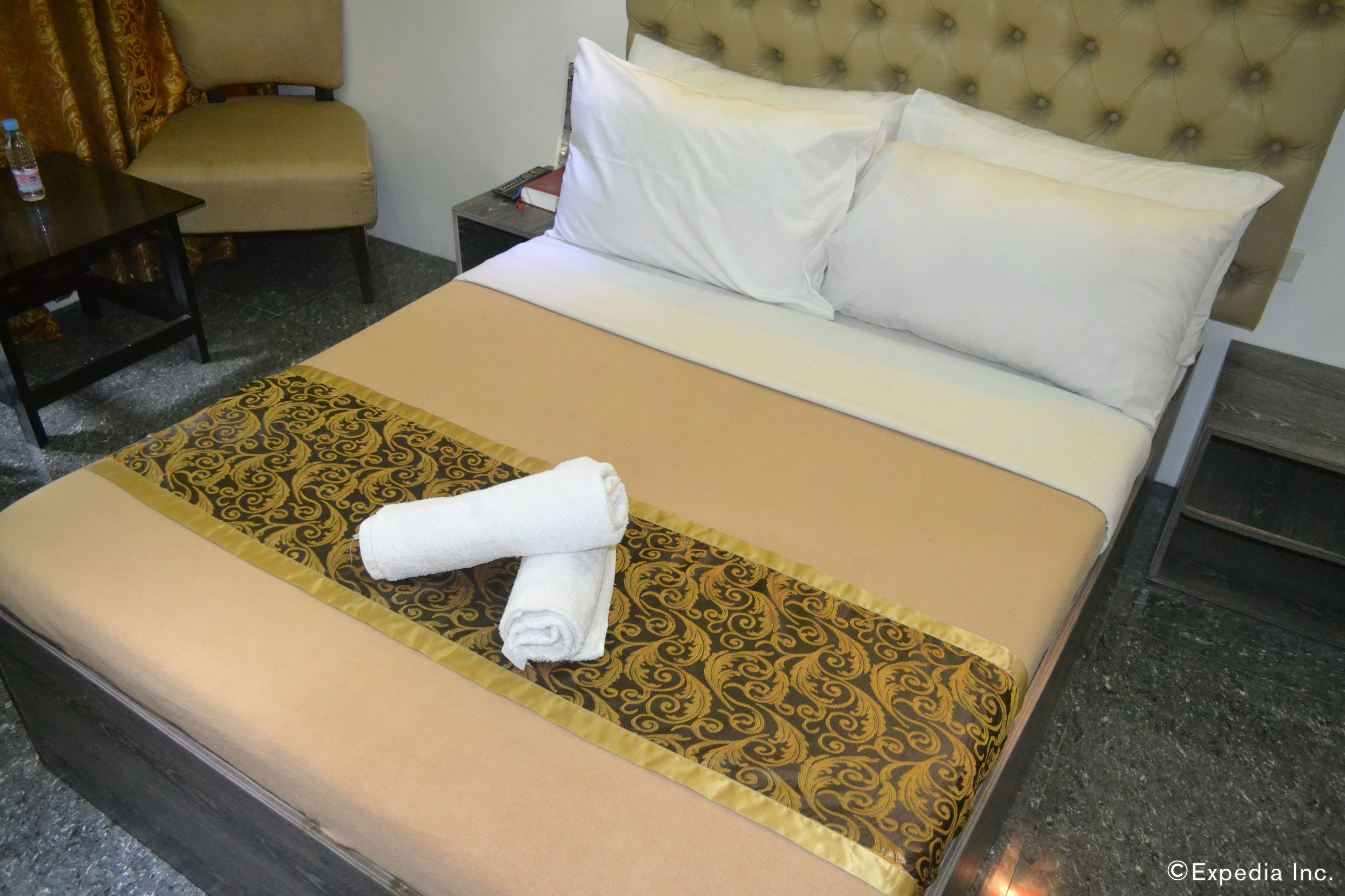 Una cama bien tendida con una colcha estampada, un par de toallas enrolladas y un cabecero con diseño acolchado.