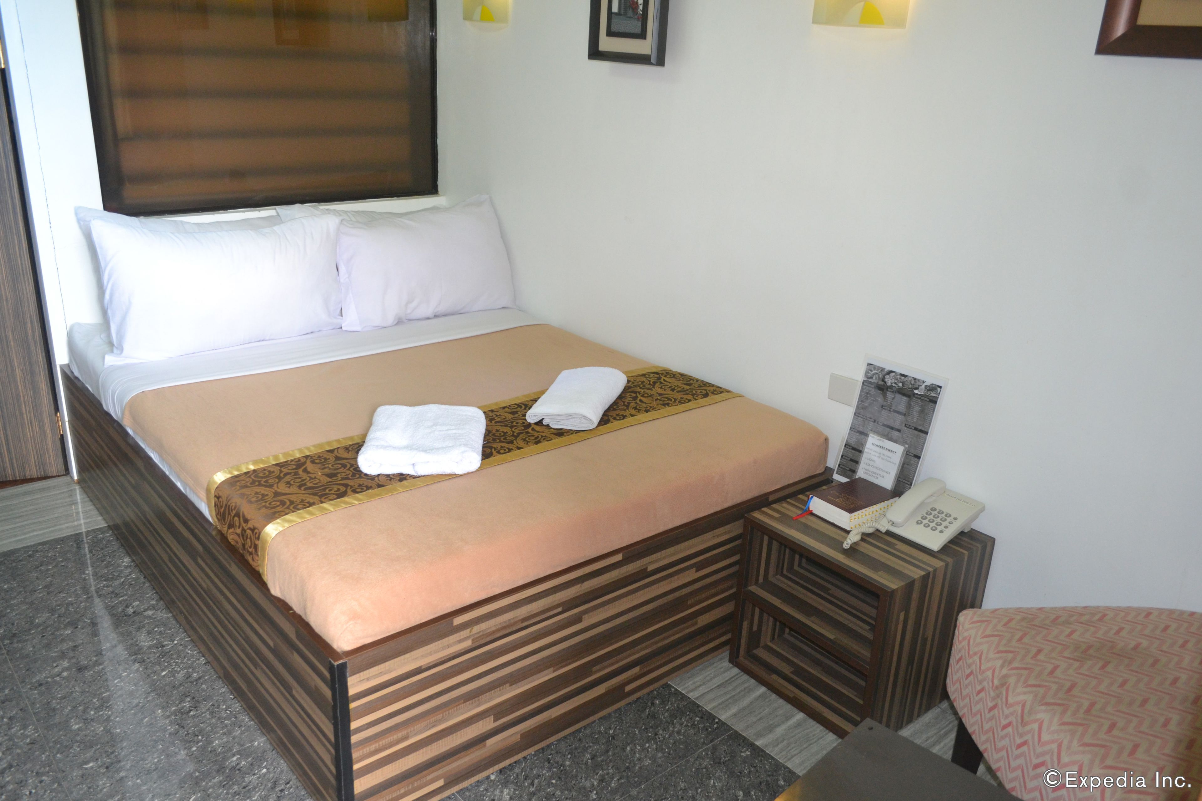 A neatly made bed with a beige bedspread and white pillows, a bedside table with a telephone and a magazine, and a small wooden nightstand.