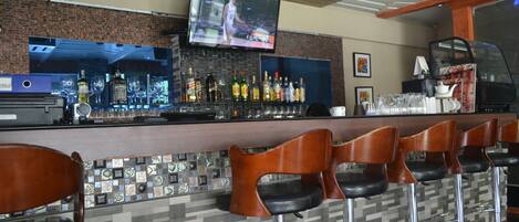 A bar area with a television mounted on the wall, a variety of bottles and glasses on the counter, and several bar stools.