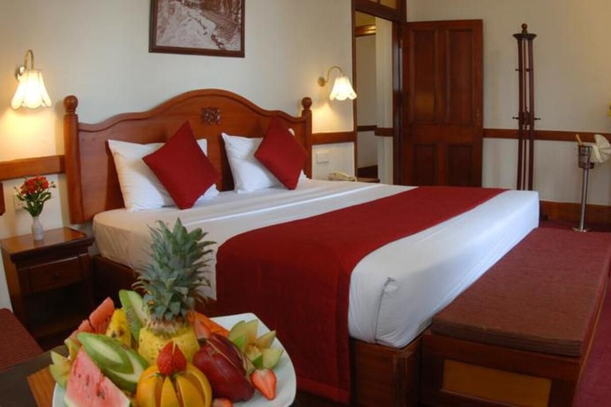 Ein Hotelzimmer mit einem großen Bett, einem Holz-Kopfstück, roter Bettwäsche, einem Obstkorb auf dem Nachttisch und einem Schminktisch mit Spiegel.