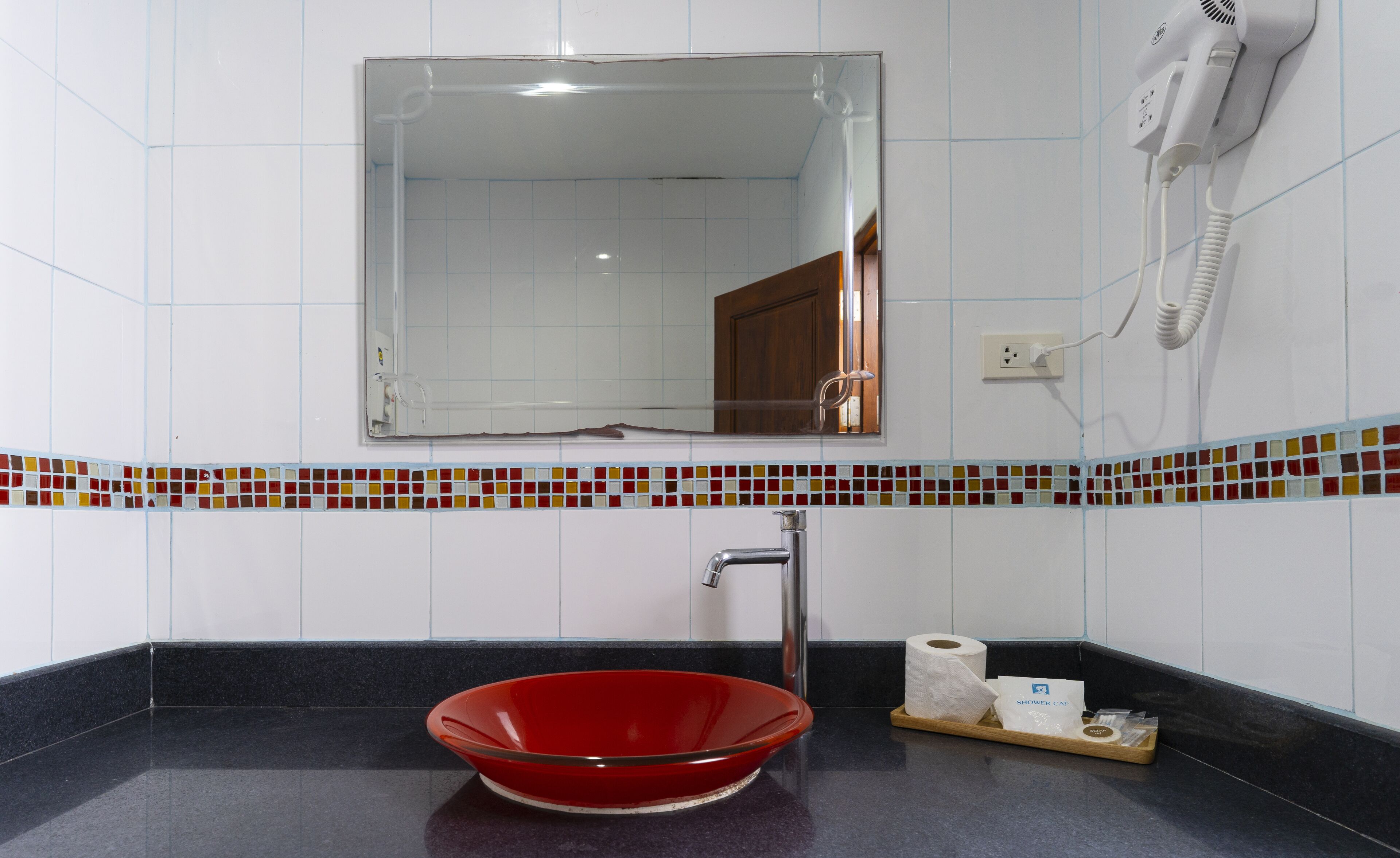 A bathroom with a red bowl sink, a mirror, a hairdryer, and a shower cap on the counter.