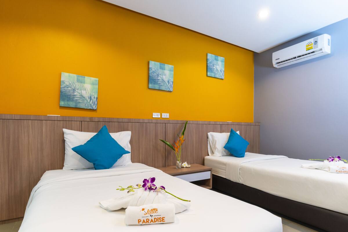 A hotel room with two beds, each with white linens and blue pillows, a wooden headboard, and a wall-mounted air conditioner.