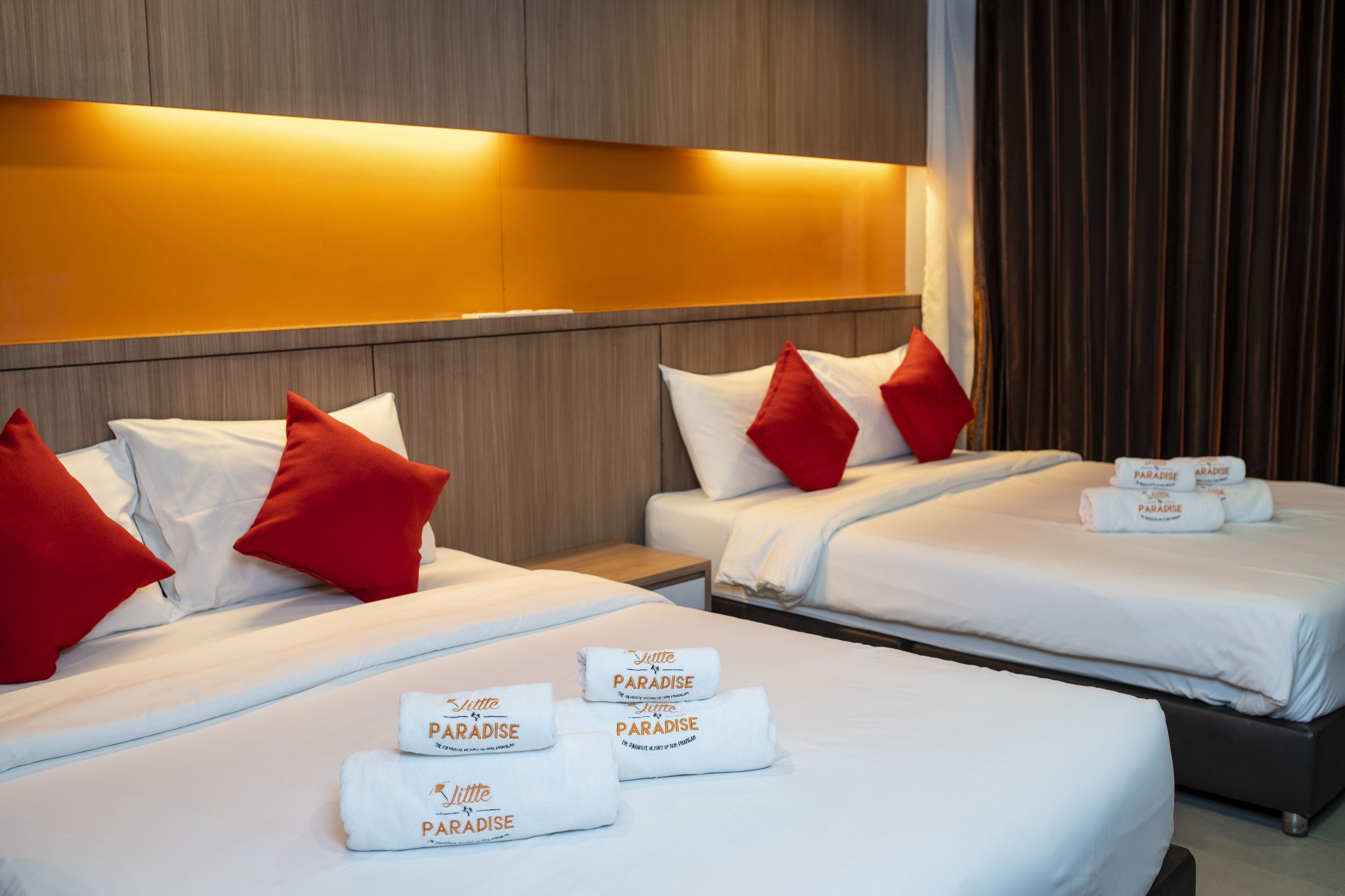 Une chambre d’hôtel avec deux lits, chacun agrémenté de draps blancs et d’oreillers rouges. On trouve des serviettes portant la marque « Paradise » sur les lits.
