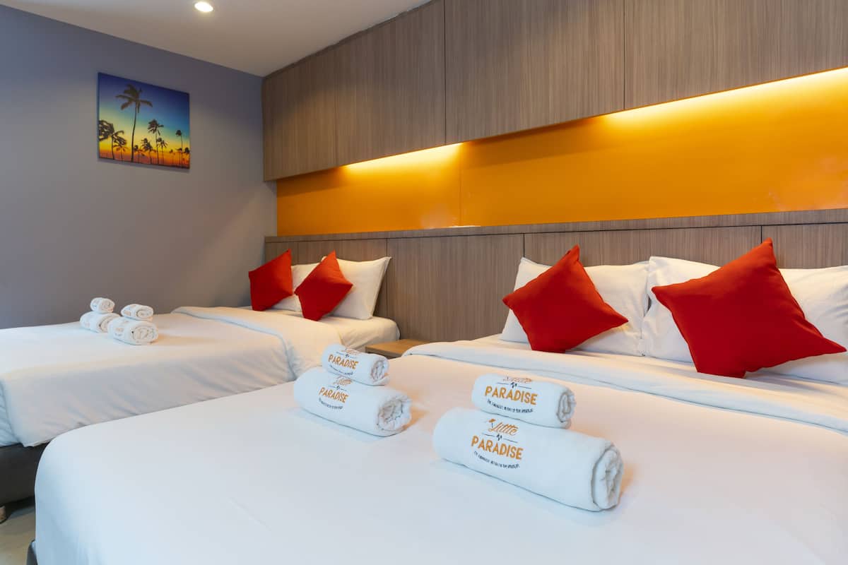 A hotel room with two beds, each with white linens and red pillows. There are rolled towels with 'Paradise' branding on the beds.