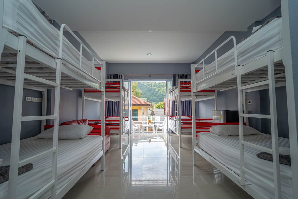A dormitory room with bunk beds, a small table, and a view of a house and trees outside.