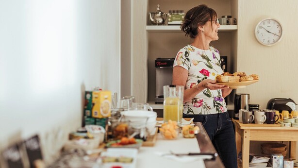 Uma pessoa segurando uma bandeja de doces em uma cozinha, com um relógio na parede, uma cafeteira e diversos utensílios na bancada.