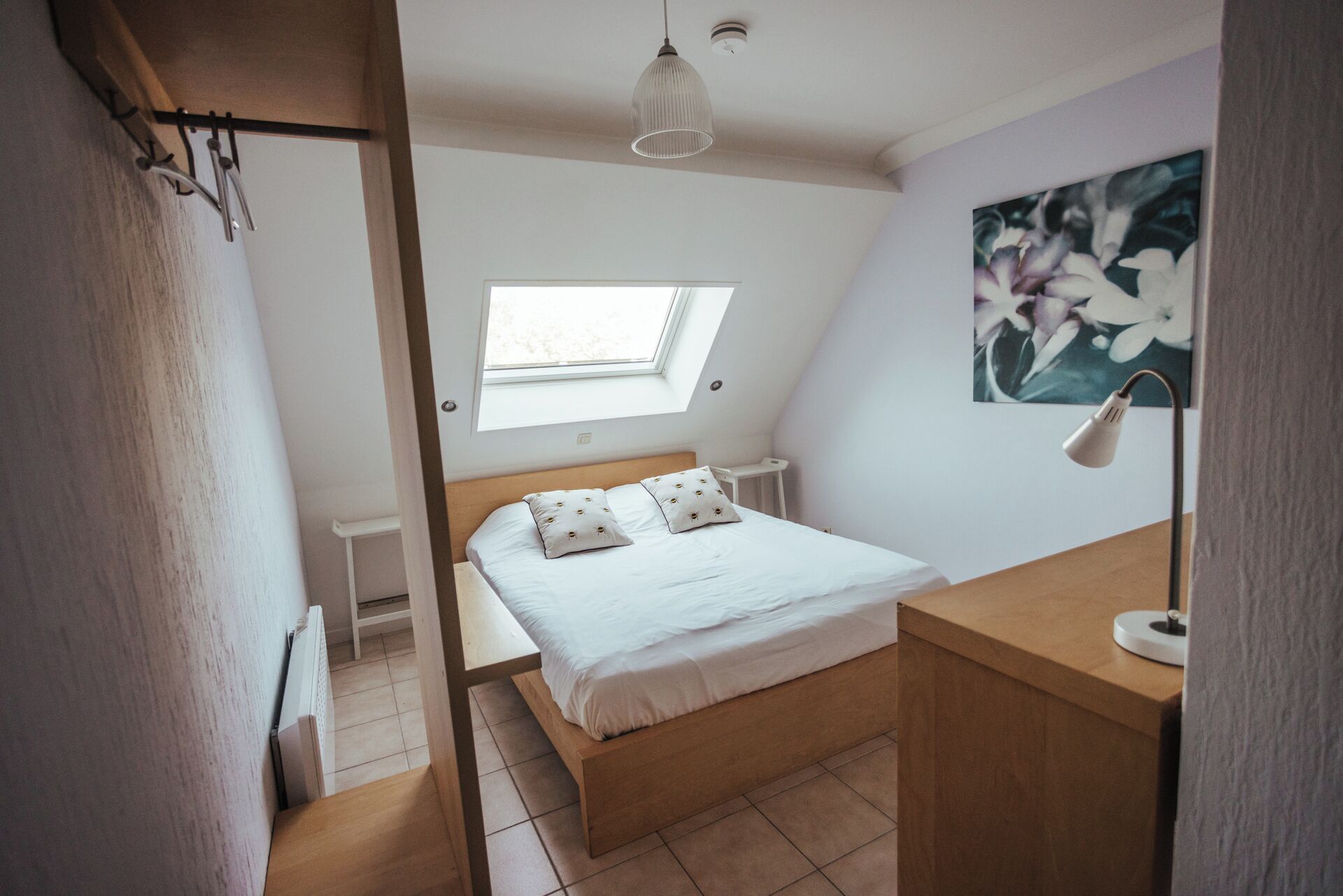 A bedroom with a bed, a desk, a lamp, a painting, and a skylight.