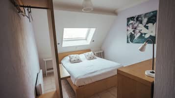 A bedroom with a bed, a desk, a lamp, a painting, and a skylight.