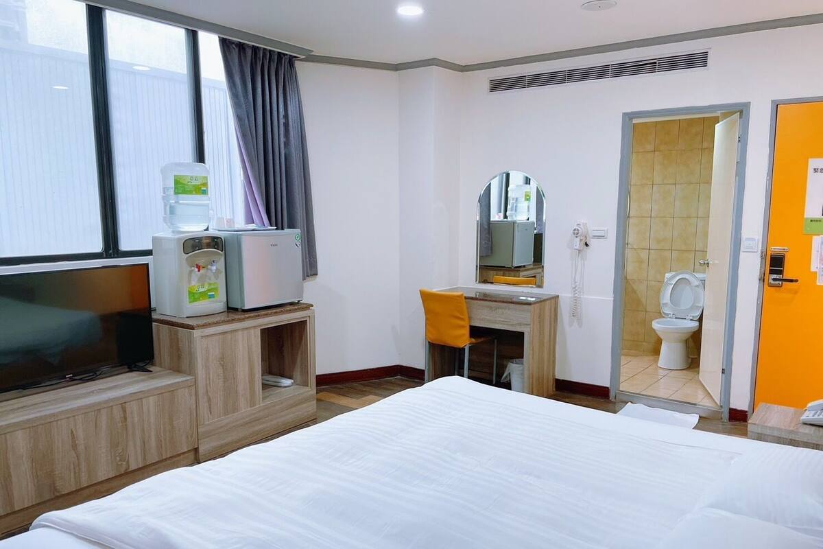 A hotel room with a bed, a desk with a chair, a television, a water dispenser, and a bathroom visible through an open door.
