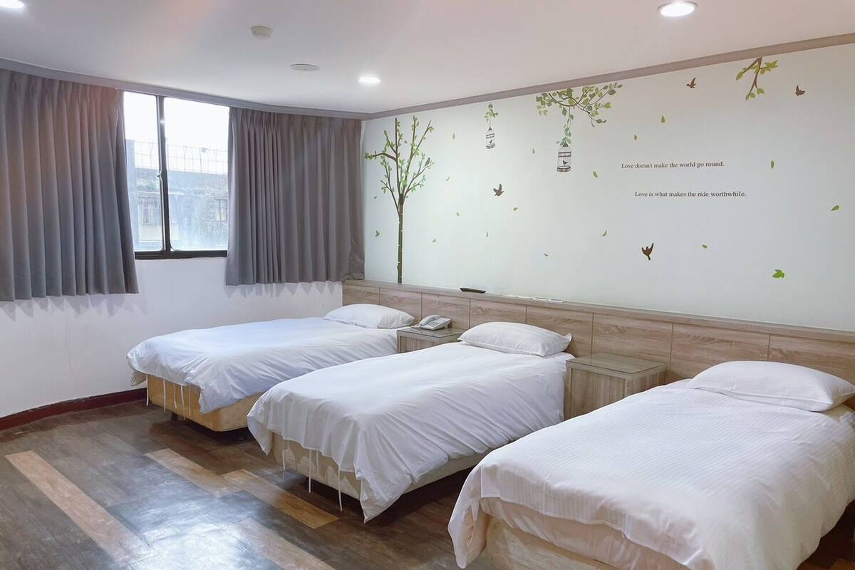 A hotel room with three single beds, a small bedside table with a phone, a window with curtains, and a wall with decorative stickers and a quote.