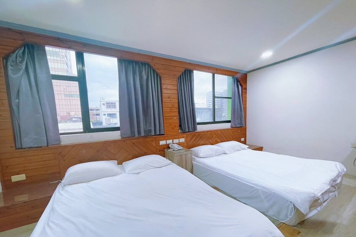 A hotel room with two single beds, wooden paneling on the walls, large windows with curtains, and a visible cityscape view.