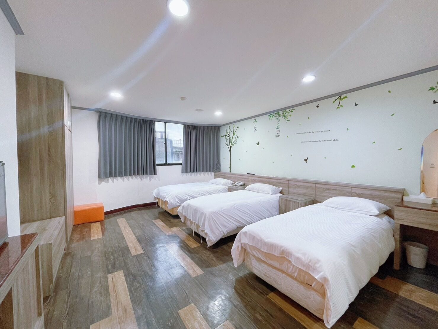 A hotel room with three beds, a wooden floor, a window with curtains, and a wall with a nature-themed design.