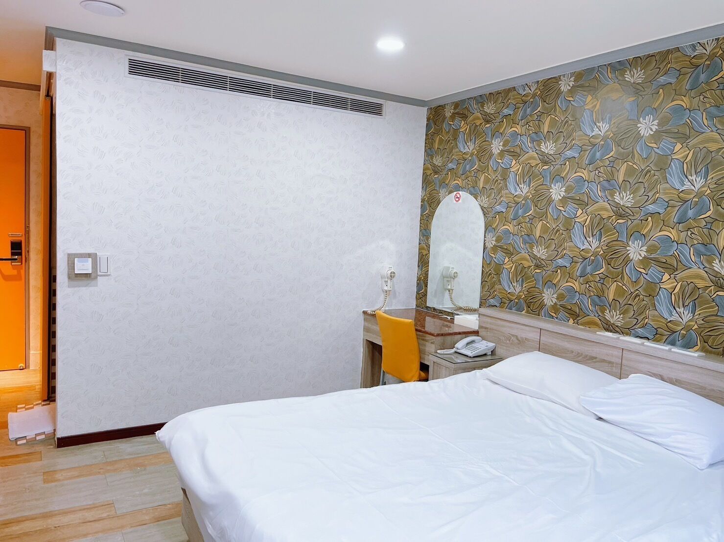 A hotel room with a bed, a bedside table with a phone, a mirror, and a floral wallpaper.